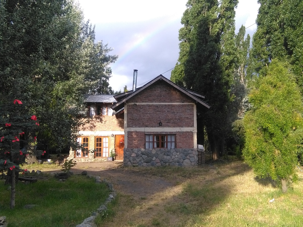 The exterior of the hostel is displayed, featuring a rustic brick facade surrounded by greenery. A garden area is present, with a mix of trees and bushes. Natural light filters through clouds, creating a serene atmosphere, while a rainbow arcs softly in the sky.