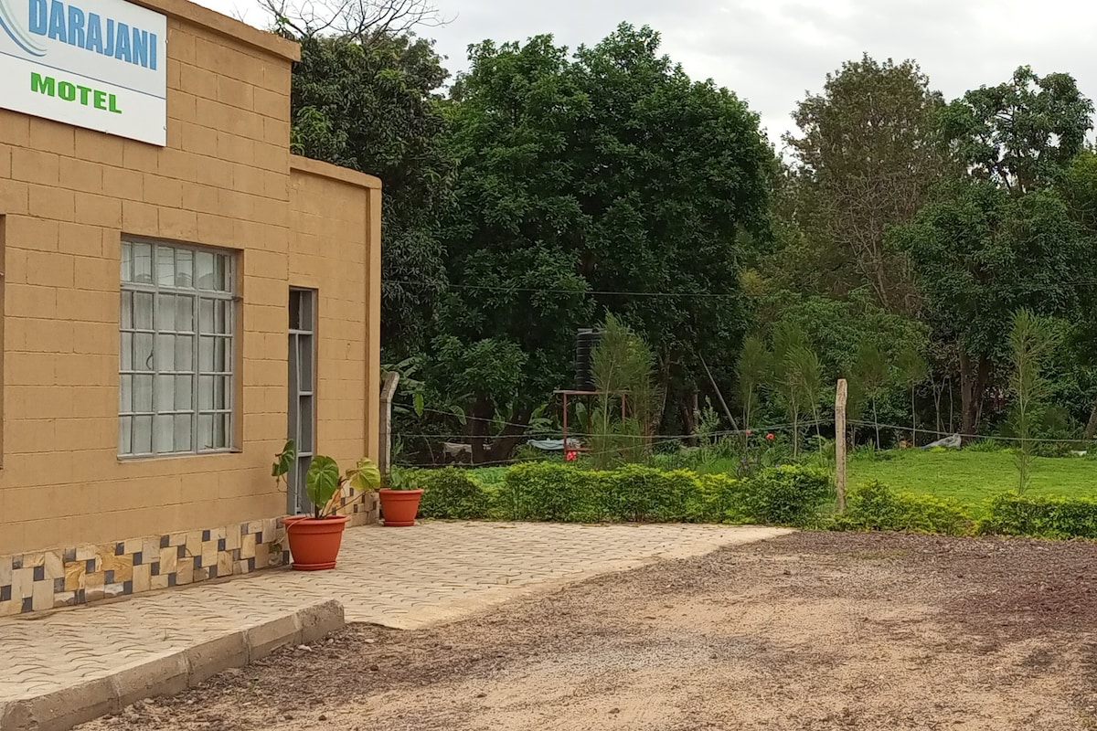 The exterior of the Darajani Motel is presented, featuring a simple building facade with large windows. Potted plants are placed near the entrance, while a spacious, gravel parking area is visible. Lush greenery surrounds the property, enhancing the natural setting.
