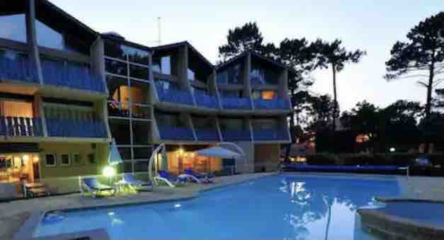 A modern hotel building features multiple balconies, illuminated in the evening light. A clear pool is situated in the foreground, with lounge chairs positioned around the water. Tall trees and a tranquil atmosphere complement the scene.