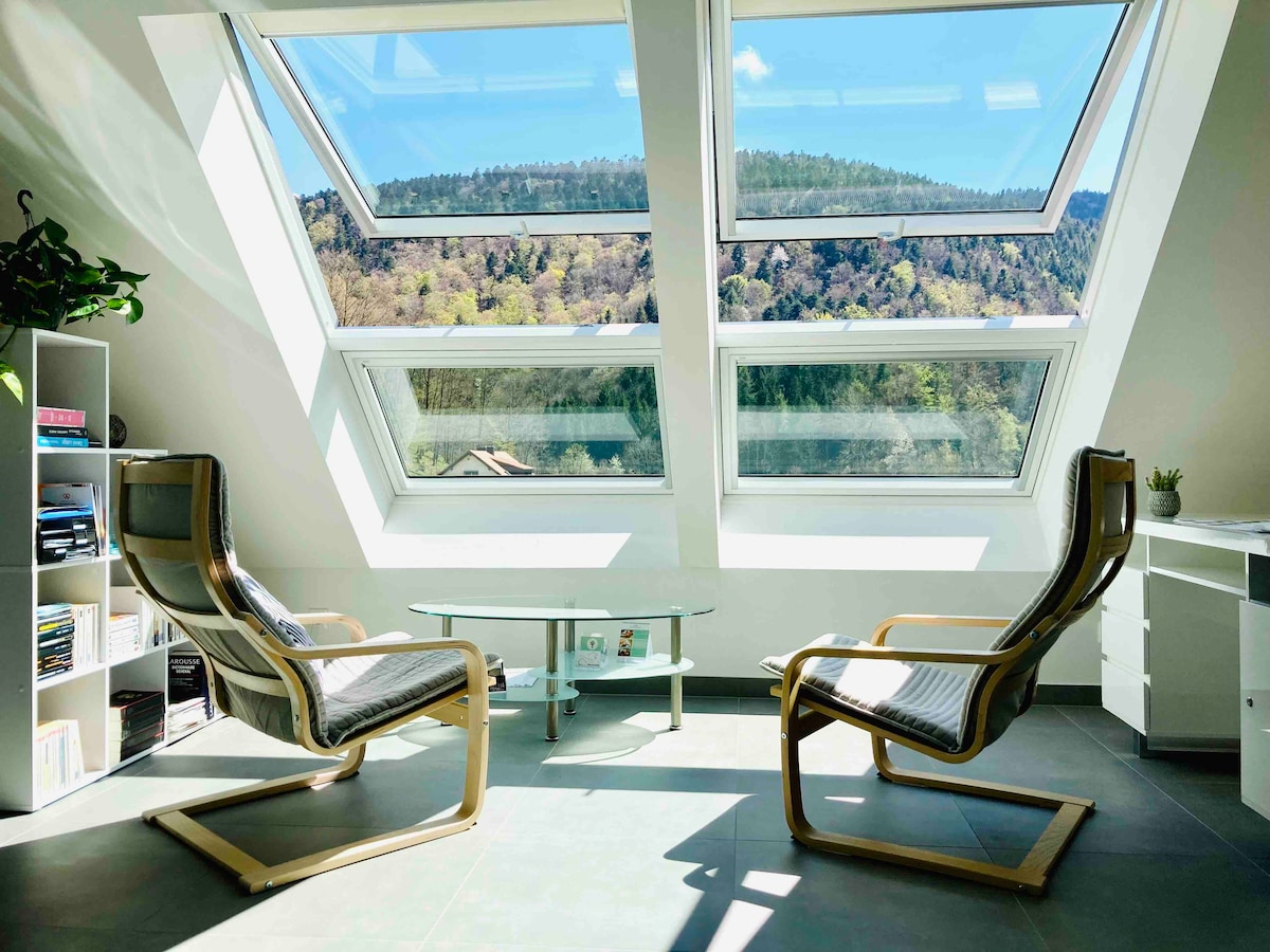 A bright corner of the living space features two wooden rocking chairs positioned beside a glass coffee table. Large skylights above provide ample natural light and offer views of the surrounding hills and trees.