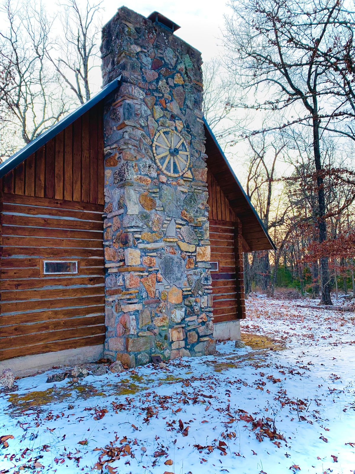 The exterior of the log cabin is showcased, featuring a prominent stone chimney adorned with a decorative wheel. Surrounding the cabin, a blanket of snow covers the ground, while the wooded area reveals leafless trees and a serene atmosphere.