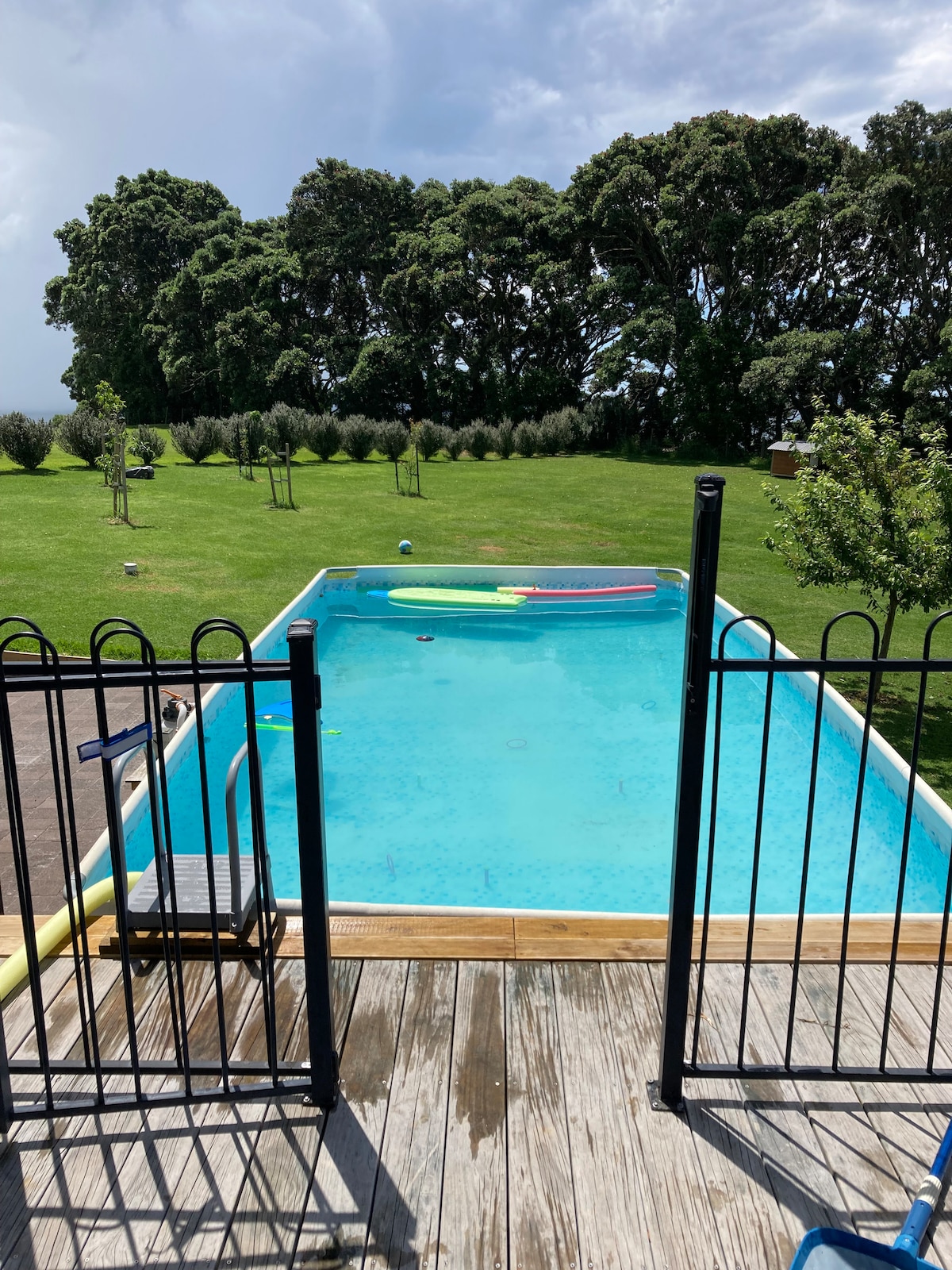 A private swimming pool is surrounded by a wooden deck and secured by a black fence. Lush greenery and trees provide a serene backdrop, while colorful pool floats rest on the clear water's surface, inviting relaxation and enjoyment.