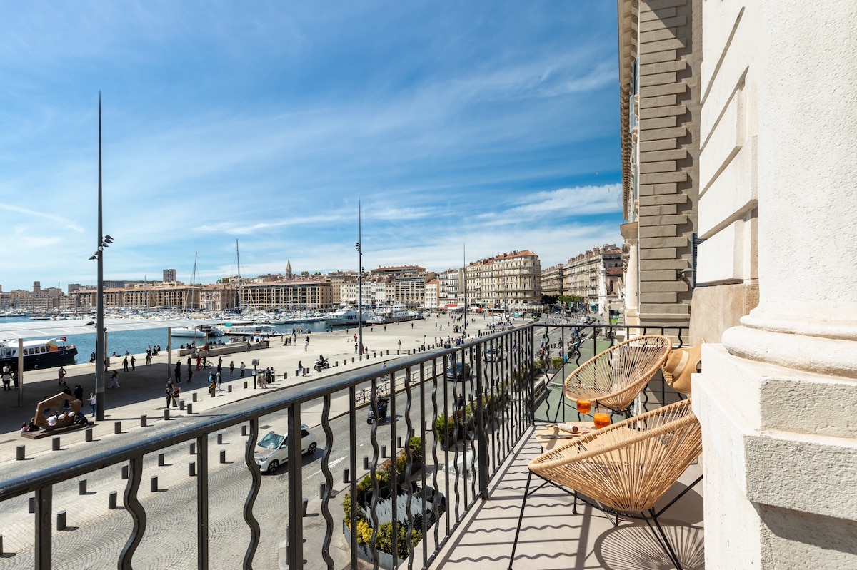 A balcony overlooks the bustling Vieux-Port, showcasing boats and lively public spaces below. Two stylish woven chairs sit invitingly on the balcony, framed by an elegant iron railing. The sky is clear, and sunlight illuminates the vibrant waterfront and nearby buildings.