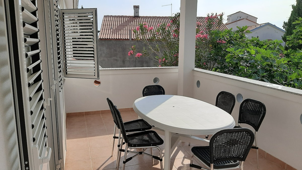 An outdoor terrace features a white round table surrounded by six black chairs. Sunlight brightens the space, with greenery visible in the background and flowers adding color. Windows with adjustable shutters allow for natural light and airflow.
