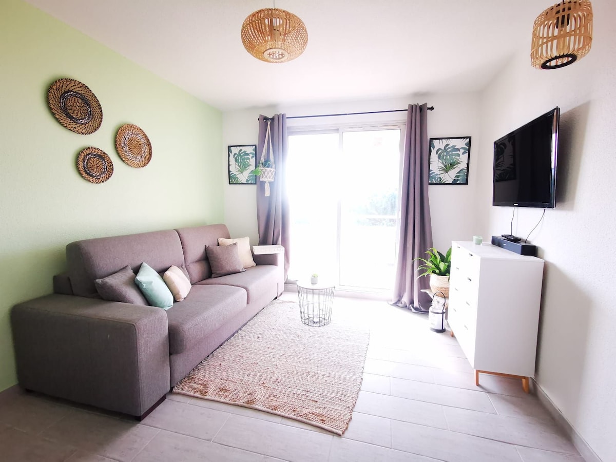 A living area is presented with a gray sofa adorned with decorative pillows. Wall art featuring natural motifs complements the light green accent wall. A television is mounted above a white console, and large windows allow natural light to fill the space, accompanied by indoor greenery.