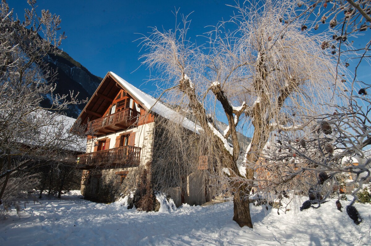 A stone and wood chalet is surrounded by a snowy landscape, with frost-covered trees nearby. The structure features a large balcony on the upper level, adding charm to the winter scene, while the snow blankets the garden area, creating a serene atmosphere.