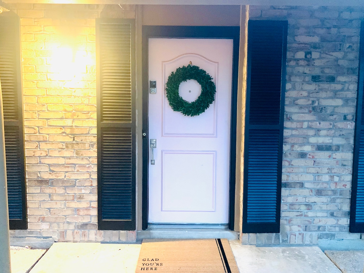 A welcoming front door is framed by dark wooden shutters and features a large green wreath. Soft light from a wall fixture illuminates the entrance, while a doormat with the words 'GLAD YOU'RE HERE' provides a friendly touch.