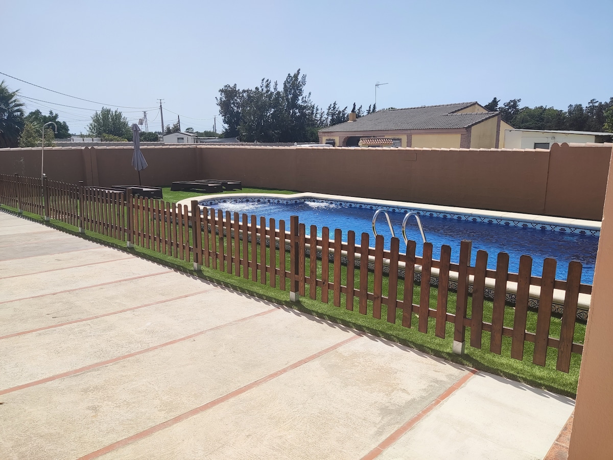 An outdoor area features a private pool surrounded by a low wooden fence. Lush green grass is visible along the edges, providing a pleasant contrast to the concrete surface. A shaded area with an umbrella can be seen in the distance, offering a spot for relaxation.