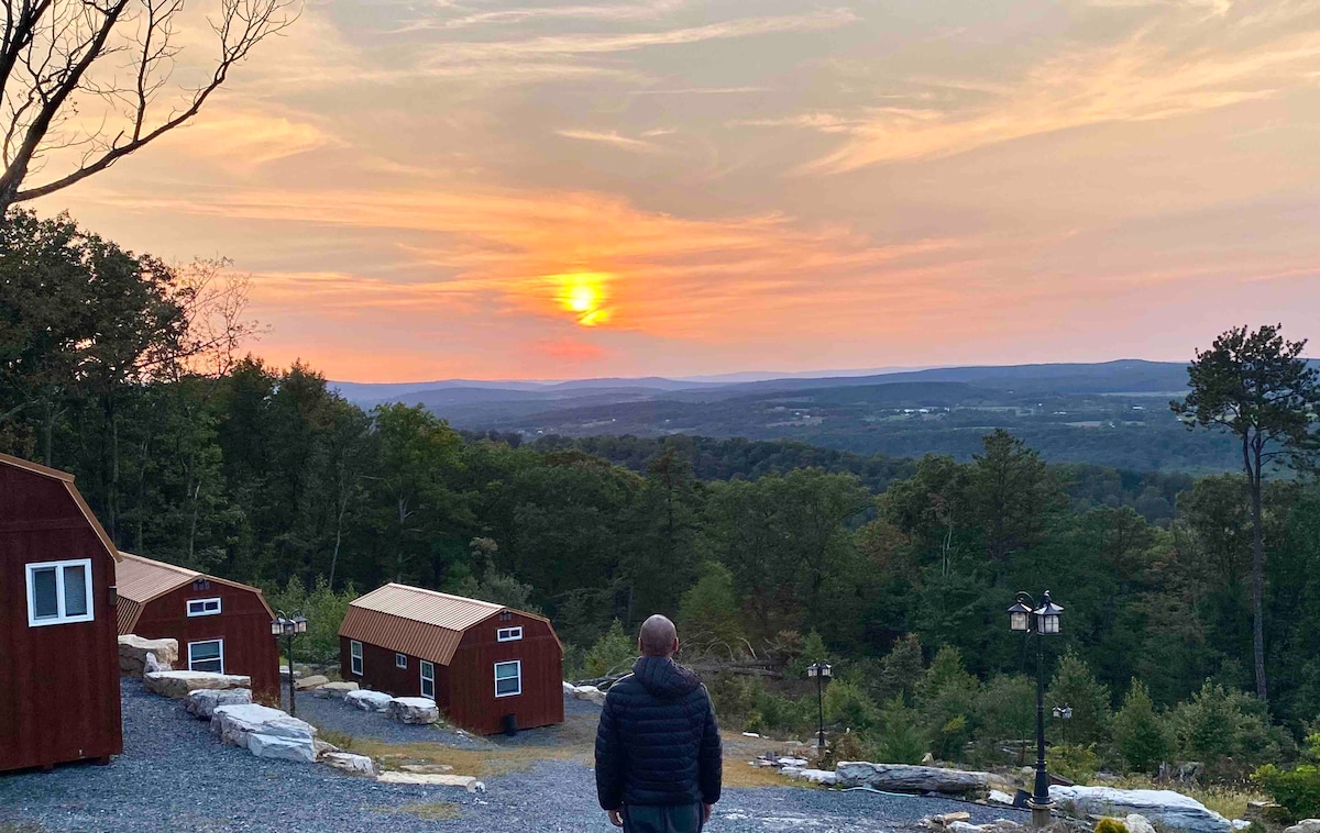 A sunset casts warm hues over the landscape, showcasing a serene view from a hillside. Several rustic cabins are situated in the foreground, surrounded by lush greenery. The tranquil scene invites contemplation and connection with nature.