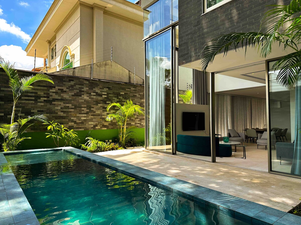 The outdoor area features a private pool surrounded by lush greenery. Large glass doors invite natural light into the living space, with a view of the pool from inside. Palm trees enhance the tropical ambiance of the setting.
