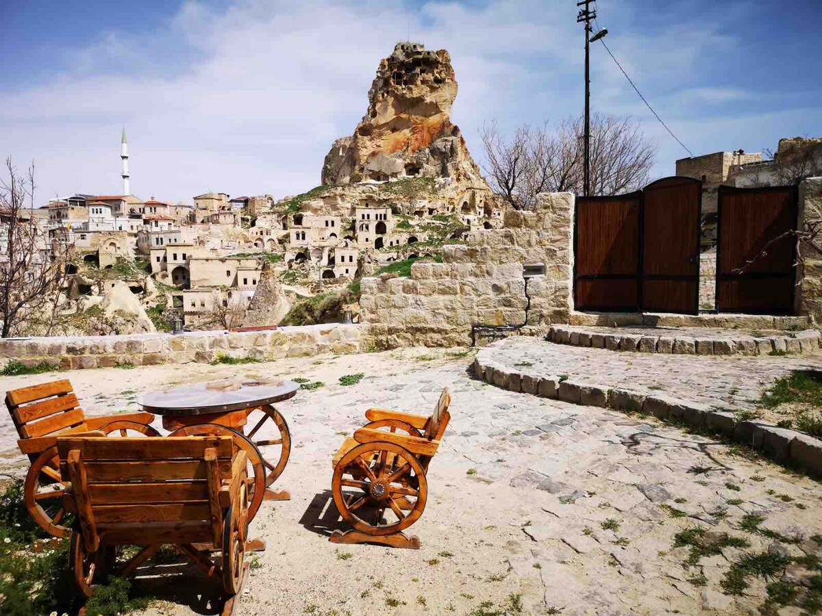 A serene outdoor seating area is depicted, featuring wooden chairs and a round table. Behind the seating, a rocky landscape is visible, with unique formations and historical structures nestled into the hillside. The sky is clear, contributing to the peaceful ambiance of the space.