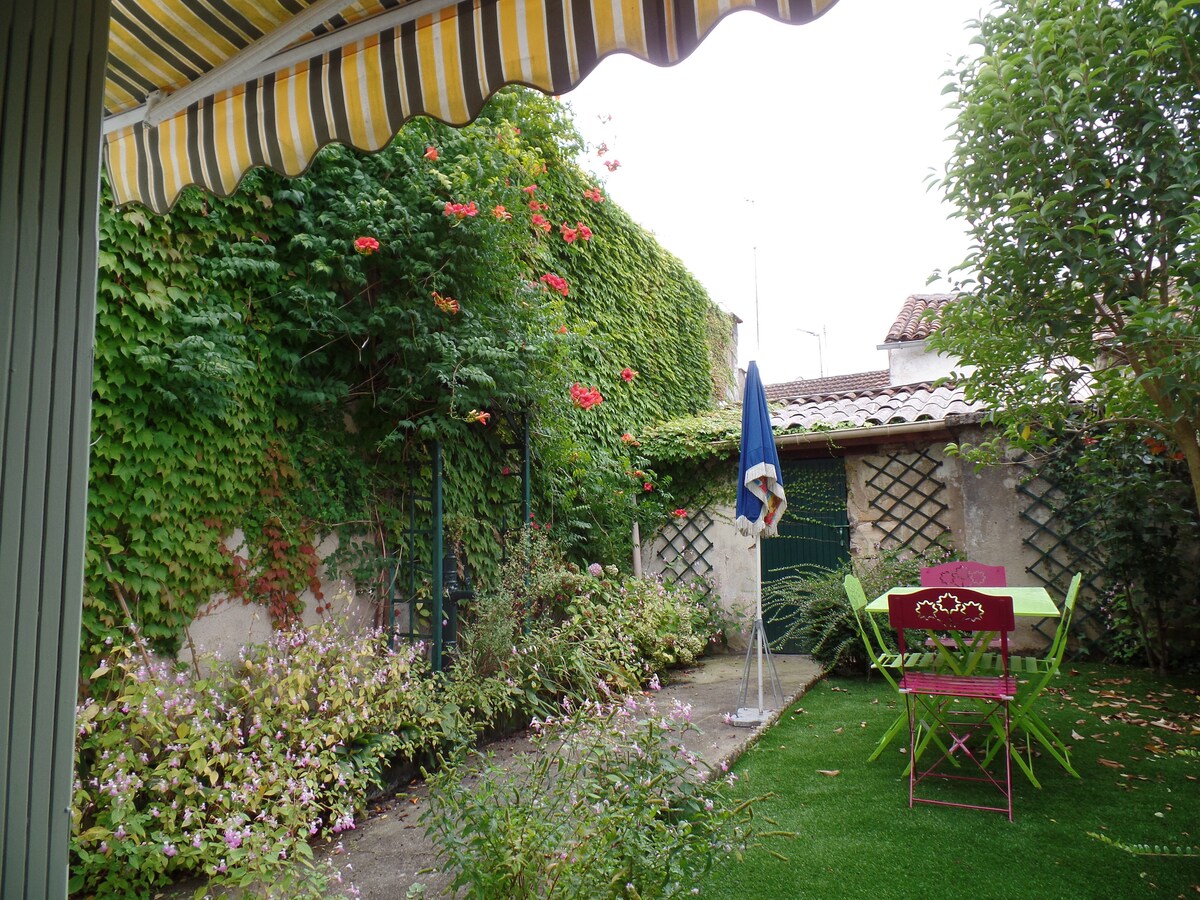 A private garden features lush greenery and colorful flowers, surrounded by ivy-covered walls. A small, round table with vibrant chairs is positioned on the manicured grass, offering a shaded area for relaxation. A large umbrella stands nearby, providing additional shade.