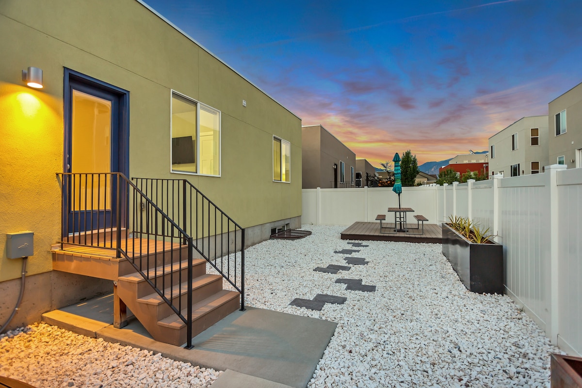 An outdoor area features a set of stairs leading to a door, surrounded by white gravel and bordered by a fenced yard. A small seating area with a picnic table can be seen in the background, under a softly illuminated twilight sky.
