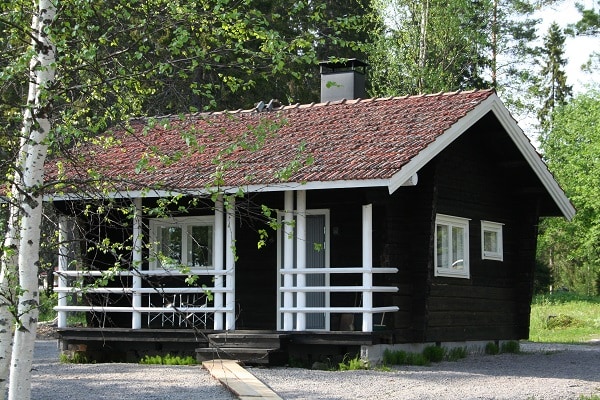 A cozy black cottage with a red-tiled roof sits amid lush greenery. The structure features multiple windows and a front deck with white railing. A wooden walkway leads to the entrance, surrounded by neatly arranged pebbles and a variety of trees.