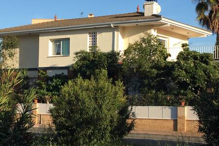 A spacious house reflects a traditional design, surrounded by lush greenery and various plants. Windows and doors are framed by vibrant foliage, while a small balcony adds to the charm. The building is positioned in a serene residential area, showcasing a clear sky above.