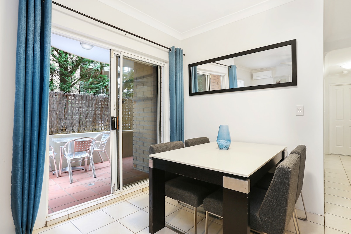 An open dining area features a modern table surrounded by four upholstered chairs. Large glass doors lead to a terrace, illuminated by natural light. Blue curtains frame the doors, and a decorative vase sits at the center of the table.