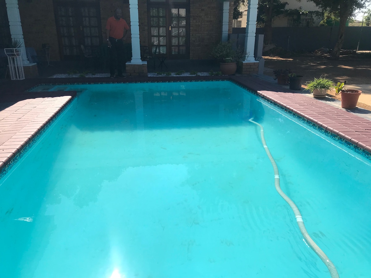 A clear swimming pool is situated in the backyard, surrounded by a patio deck. The water reflects sunlight, creating a serene ambiance. Potted plants are placed near the pool, adding a touch of greenery to the environment. A person stands near the entrance of the house.