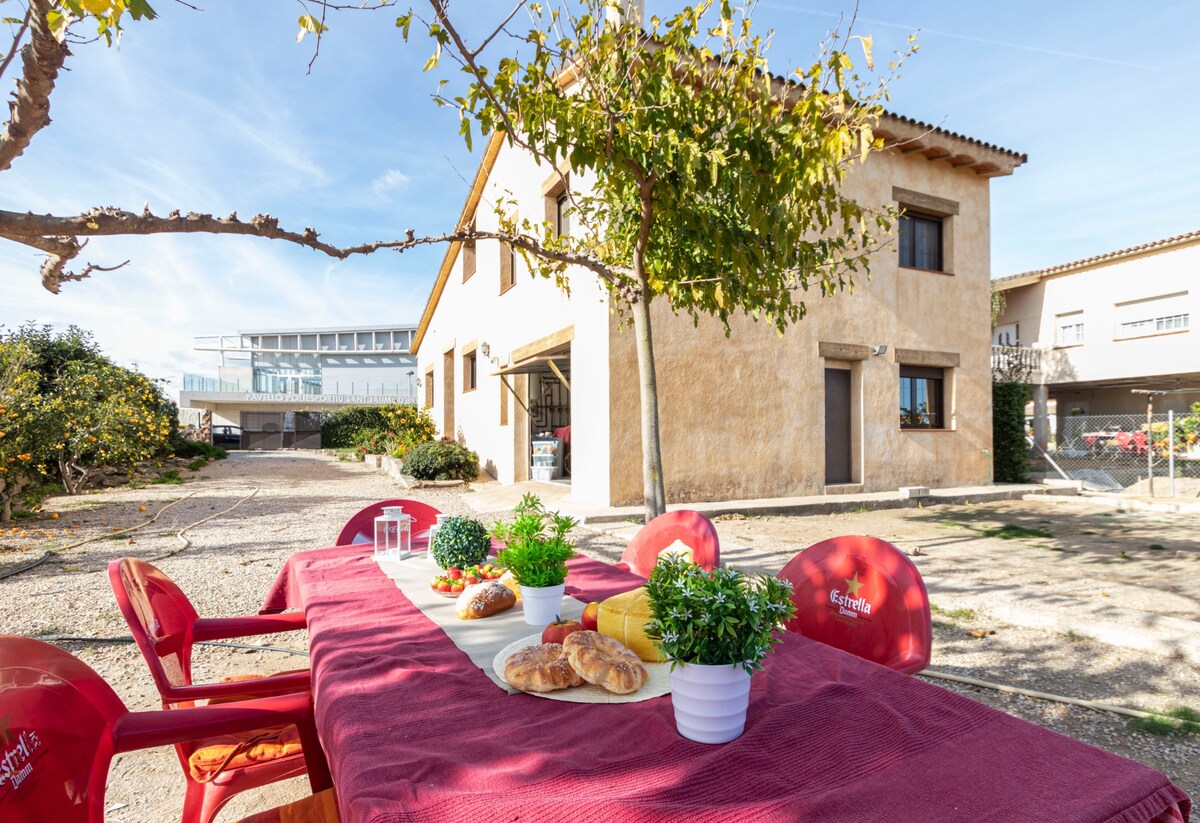 An outdoor dining area is presented with a long table adorned with fresh food and greenery. Red chairs provide seating, and the house is visible in the background, showcasing its earthy facade and windows. A tree offers shade to the gathering space.