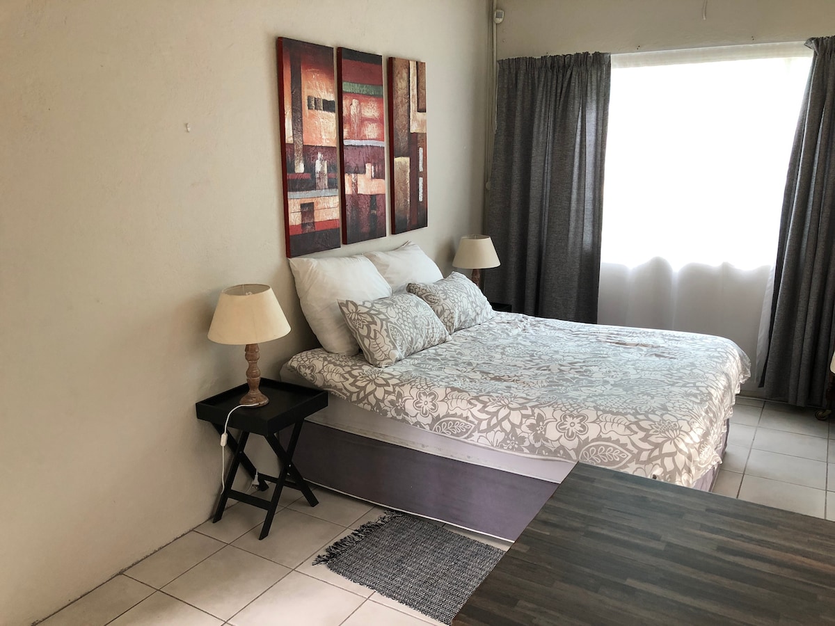 A serene bedroom features a spacious bed adorned with patterned bedding and soft pillows. Two bedside lamps provide gentle illumination. A small table sits adjacent to the bed, and curtains frame a large window allowing natural light to enter the room.