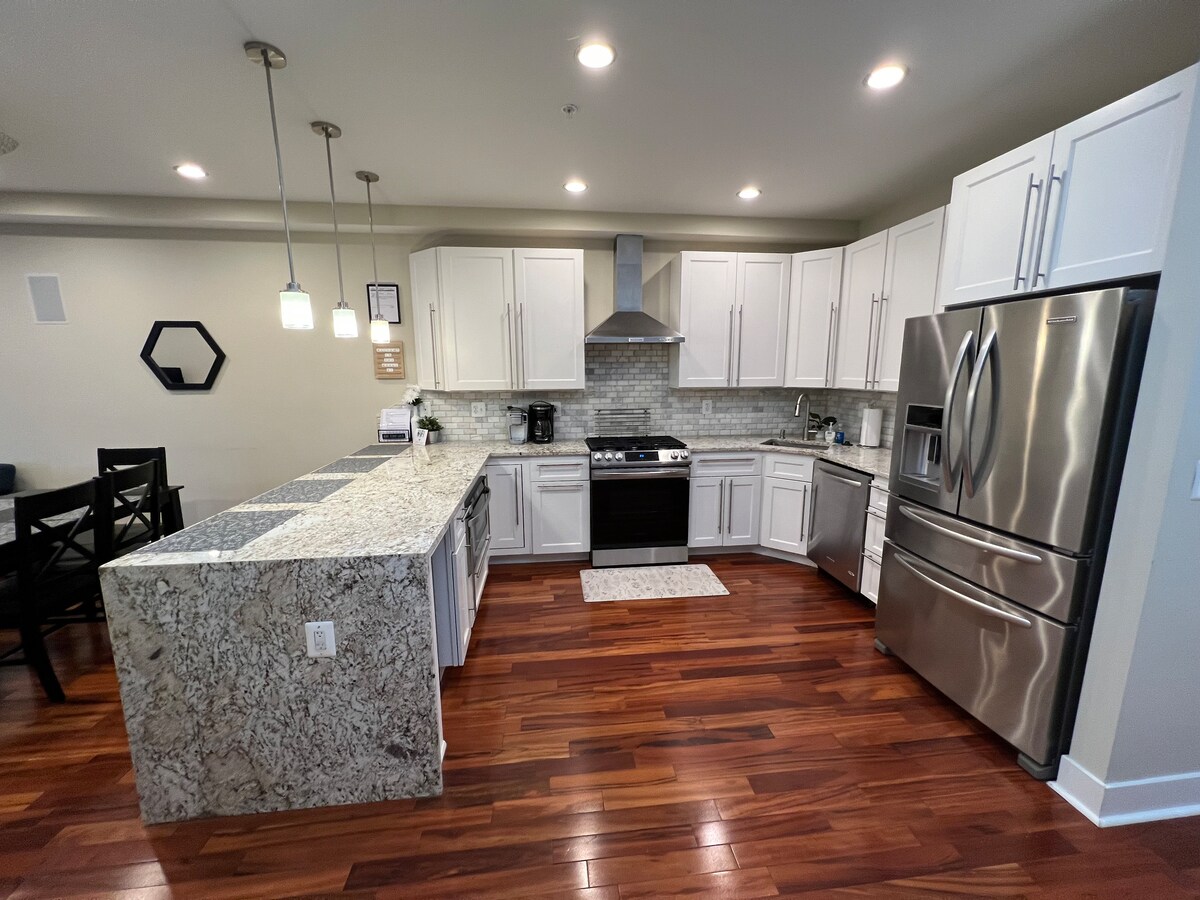 The kitchen features modern stainless steel appliances and white cabinetry, complemented by granite countertops. Pendant lights illuminate the space, while a patterned rug adds texture to the wooden floor. A dining area is visible in the background, enhancing the open concept layout.