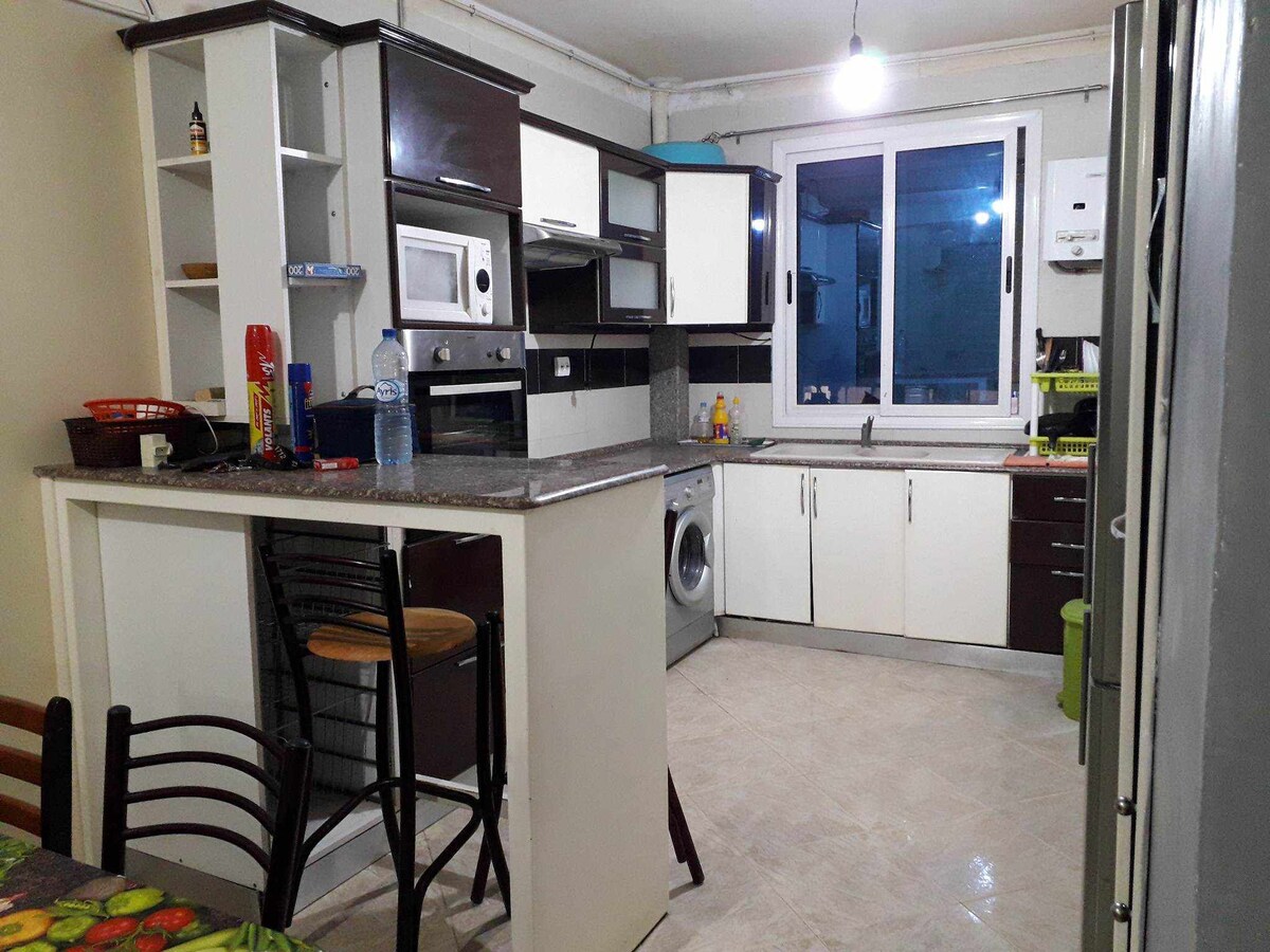 The kitchen features sleek cabinetry and a spacious countertop, equipped with modern appliances including a microwave and washer. A window allows natural light to brighten the space, complemented by a dining area with wooden chairs and a round table.