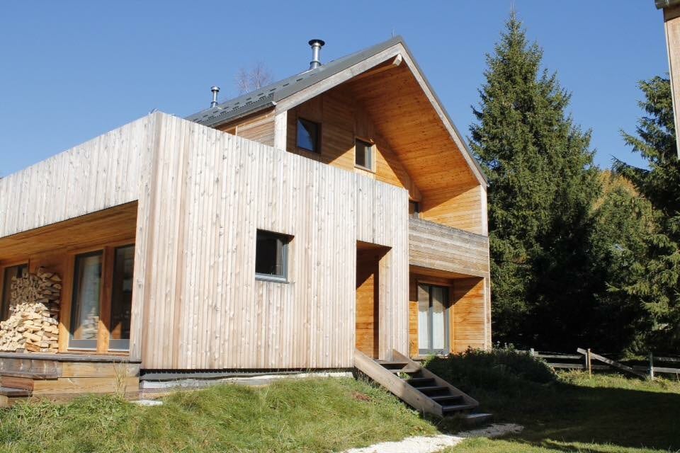 A modern wooden house is presented, featuring a combination of natural wood siding and large windows. The structure includes a sloping roof and a ramp leading to the entrance. Surrounding greenery and trees enhance the natural setting.