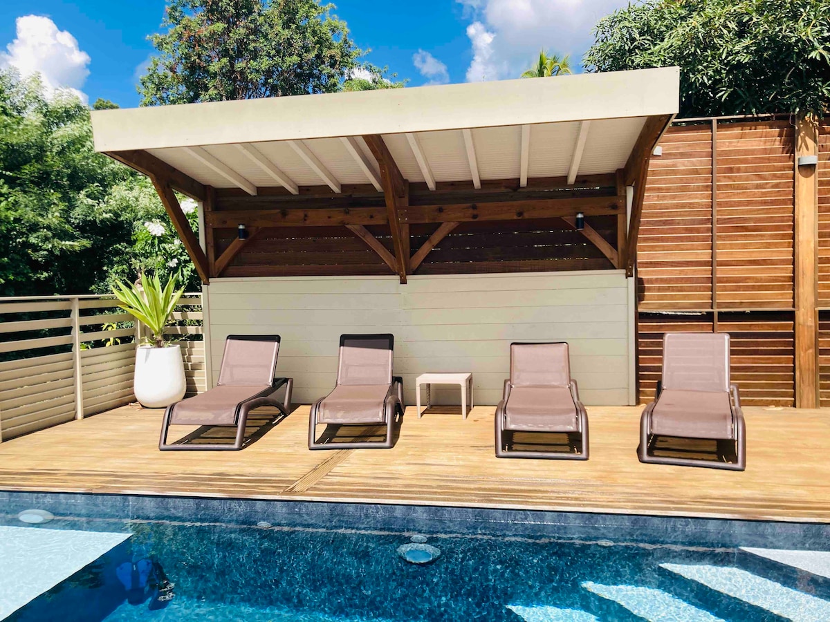 A poolside area features four lounge chairs arranged neatly on a wooden deck. The overhead structure provides shade, complemented by lush greenery in the background. The inviting pool gleams under sunlight, enhancing the tranquil setting.