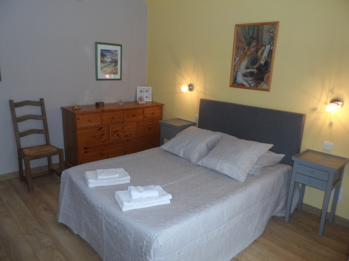 The bedroom features a spacious double bed with light bedding positioned against a soft gray wall. Two small bedside tables flank the bed, each equipped with modern wall sconces. A natural wood dresser is positioned against the opposite wall, complemented by a piece of framed artwork.