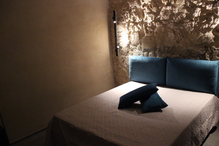 A cozy sleeping area features a spacious bed dressed in a light quilt. Two teal cushions are positioned neatly on the bed. The stone wall offers a textured backdrop, complemented by soft lighting mounted on the wall.