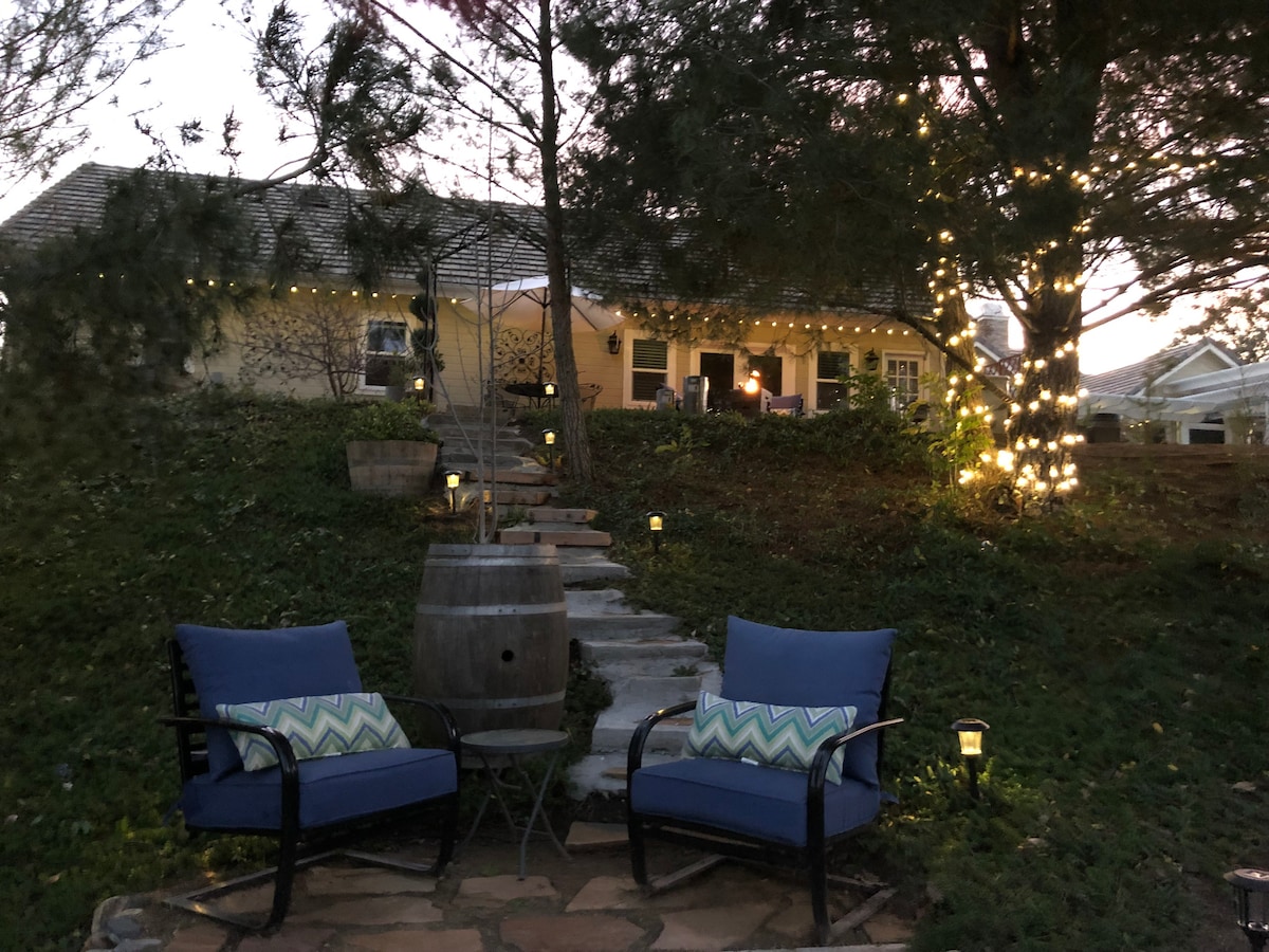 A serene outdoor space features two blue chairs adorned with a patterned cushion, positioned by a stone path leading to the inviting entrance of the guest suite. Soft lighting illuminates the surroundings, enhancing the tranquil atmosphere amidst greenery and trees.