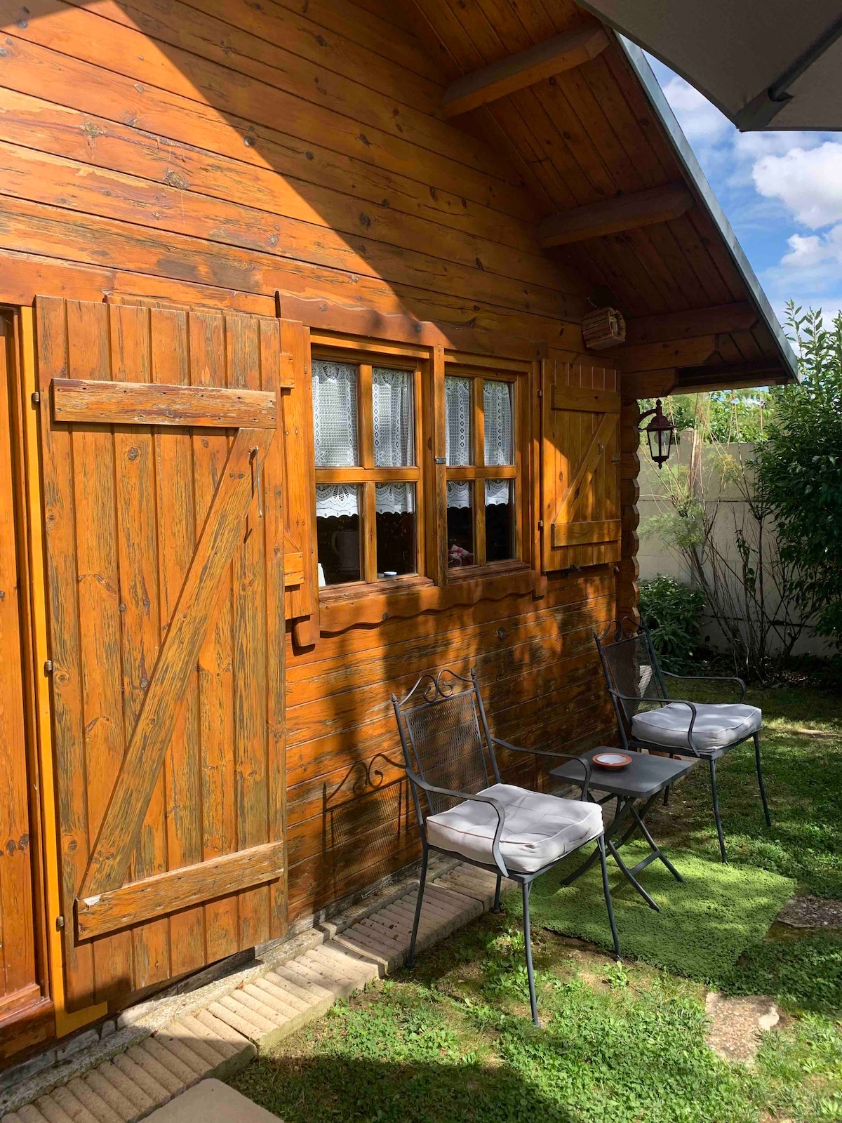 The exterior of the wooden chalet displays charming shutters beside square windows. A small seating area featuring three metal chairs is positioned on a grassy patch, inviting relaxation. Sunlight filters through the surrounding greenery, enhancing the serene setting.