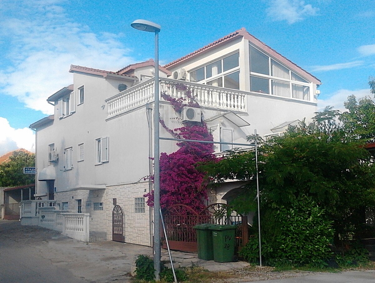 A two-story villa is presented, featuring a white exterior complemented by vibrant flowering plants. A balcony with a railing is visible on the upper level, while large windows allow natural light to fill the interior. Surrounding greenery and a lamppost add to the scene.