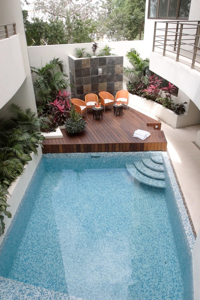 A serene swimming pool is surrounded by lush greenery and vibrant plants, featuring a wooden deck with two orange chairs and a small table. The pool's clear blue water invites relaxation, while natural stone accents add to the peaceful ambiance.