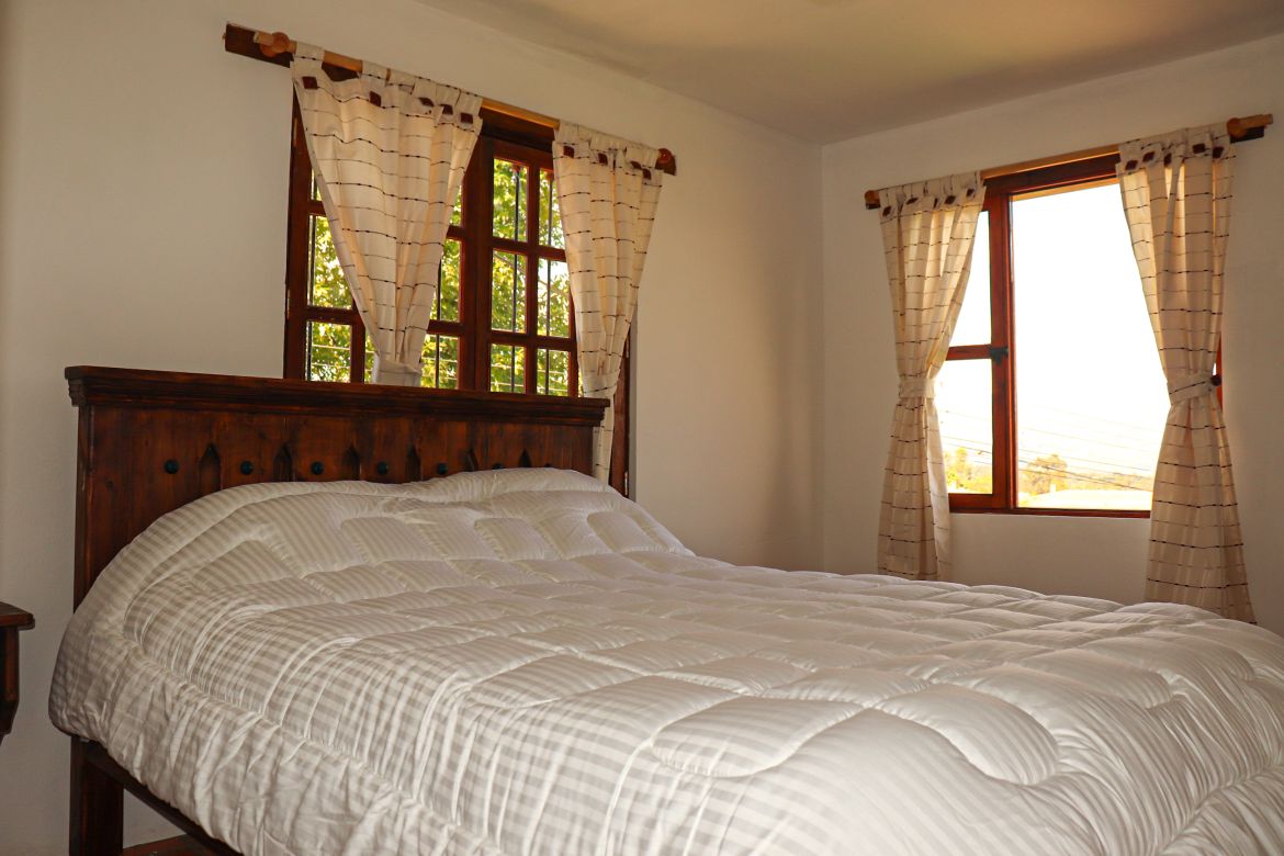 A spacious bedroom features a large bed covered with a white comforter. Natural light enters through two windows adorned with light curtains, illuminating the warm wood tones of the bed and surrounding decor.