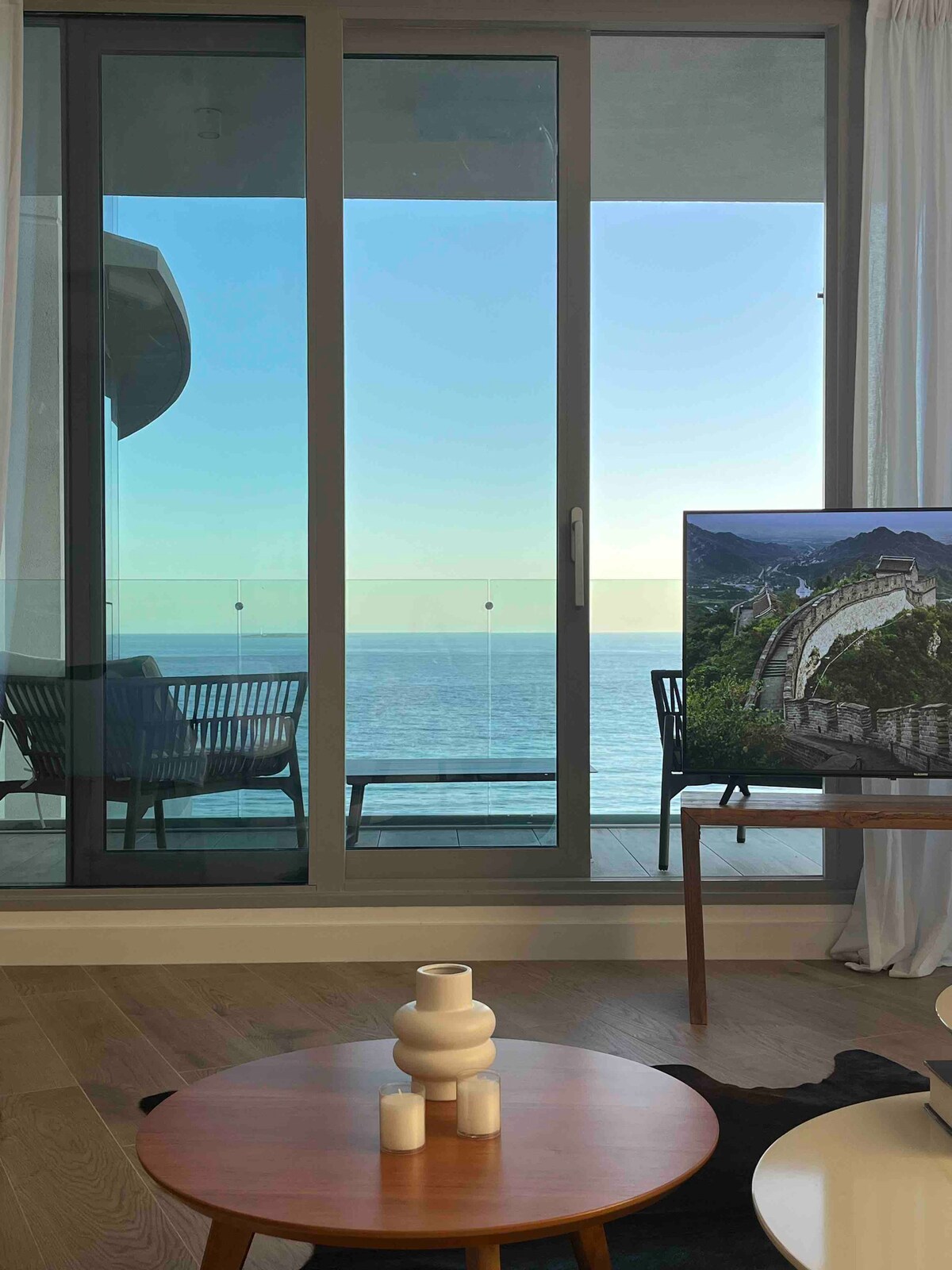 A spacious living area is showcased, featuring a low wooden coffee table and a large television. Floor-to-ceiling glass doors open to a balcony with an unobstructed view of the serene ocean, allowing natural light to fill the space.