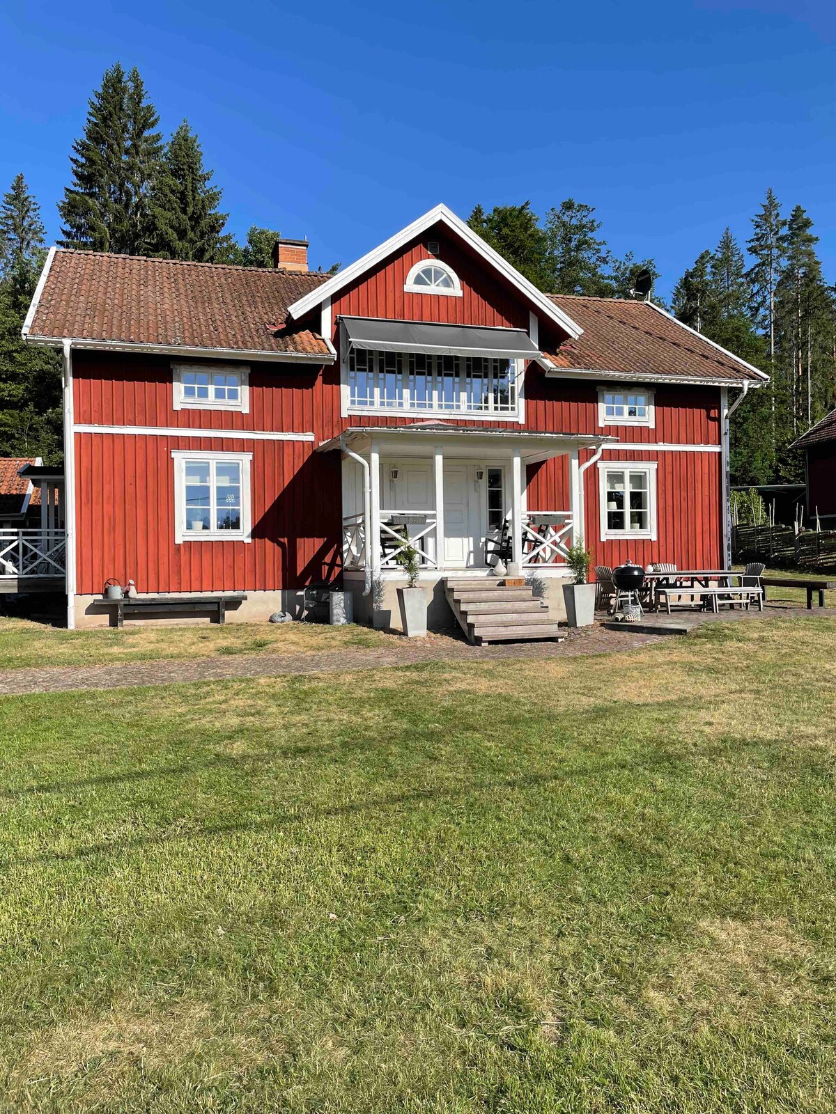 A red wooden house with a gabled roof is set against a backdrop of tall trees and blue sky. The entrance is framed by a small porch with steps leading up to it. Large windows are visible on the facade, enhancing natural light.