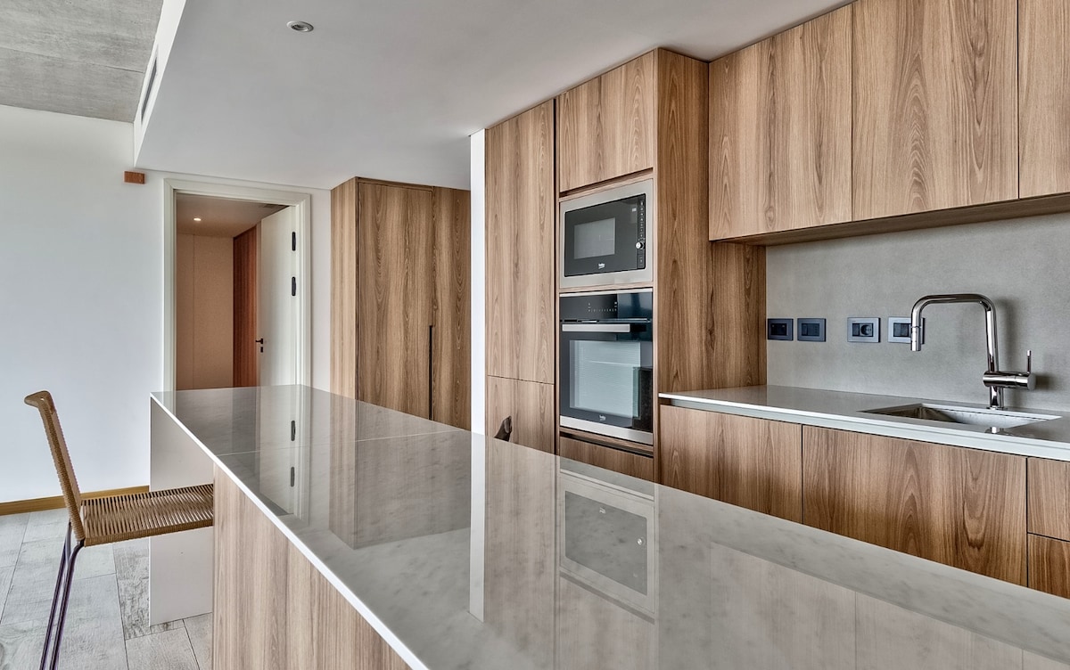 A modern kitchen area showcases sleek wooden cabinetry, complemented by a polished countertop. Built-in appliances, including an oven and microwave, are integrated seamlessly into the design. A bar-style chair stands next to the countertop, enhancing the functional layout of the space.