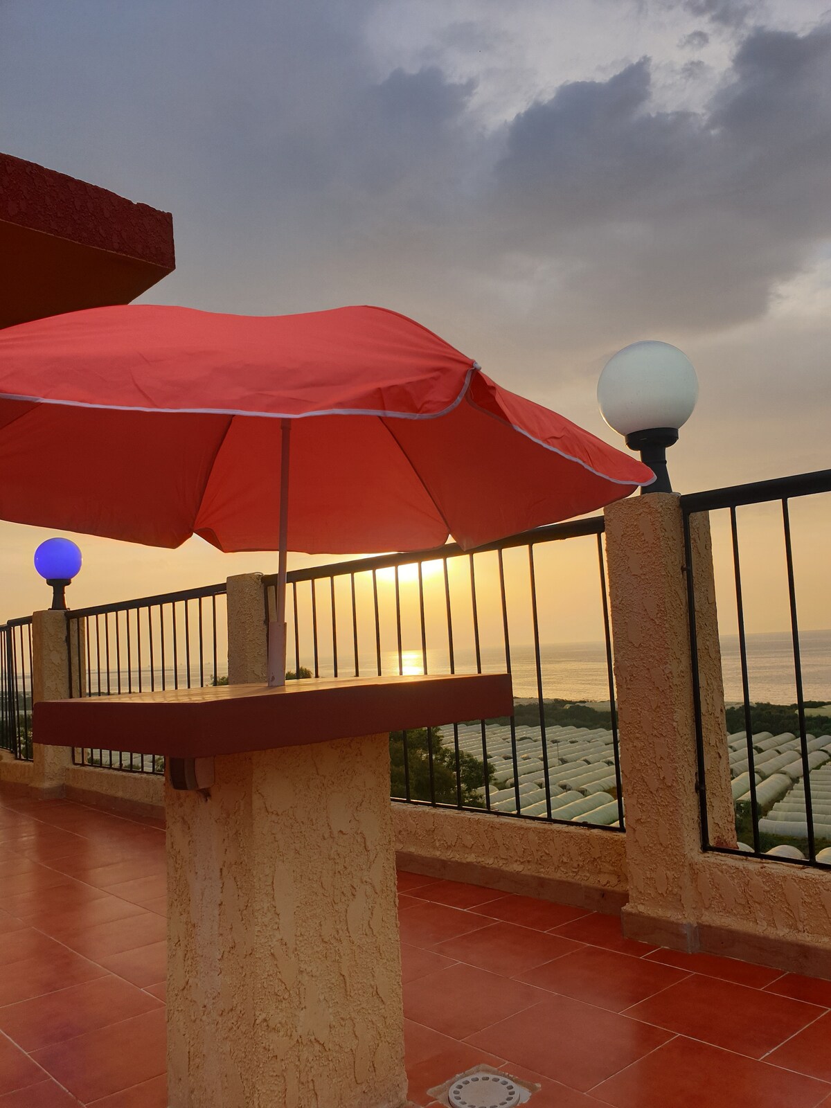 An outdoor terrace is featured, equipped with a red umbrella and gentle lighting from round lamps. The sun is seen setting over the horizon, casting warm hues across the sky and reflecting off the water, creating a serene atmosphere.