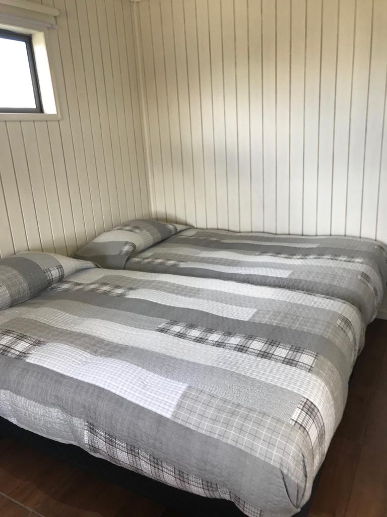 A cozy sleeping area features two spacious beds with a grid-patterned gray and white bedding. Wooden flooring complements the light-colored walls, while natural light enters through a small window.
