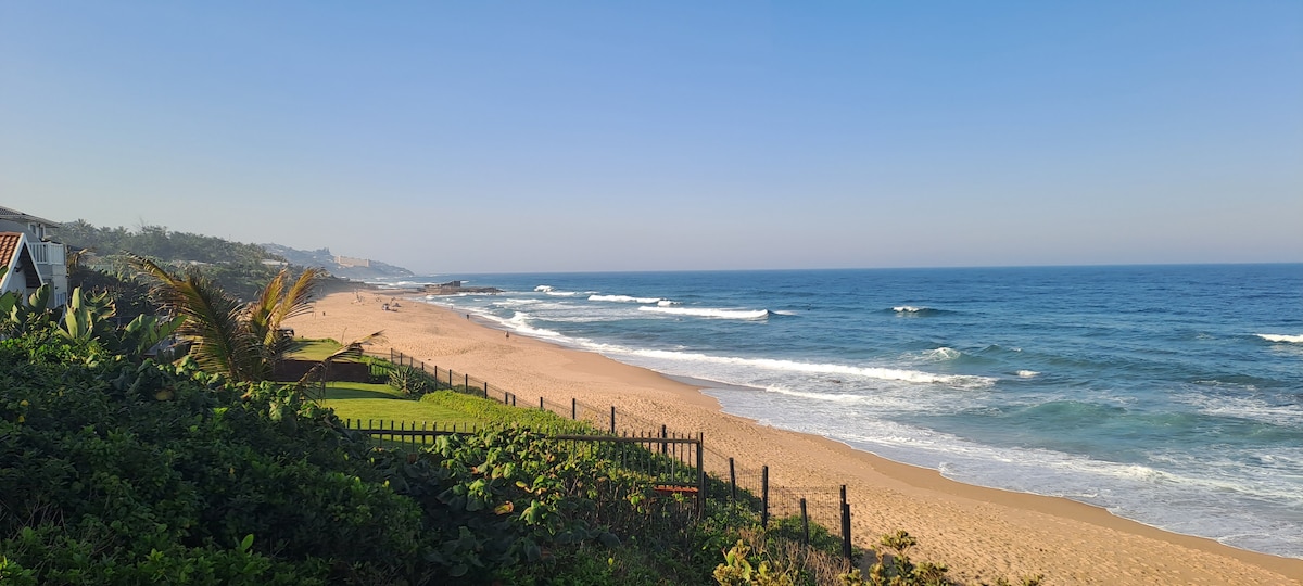 A panoramic view of an expansive sandy beach is captured with gentle waves lapping at the shore. Lush greenery borders the scene, with beachgoers visible in the distance, enjoying the tranquility of the shoreline under a clear blue sky.