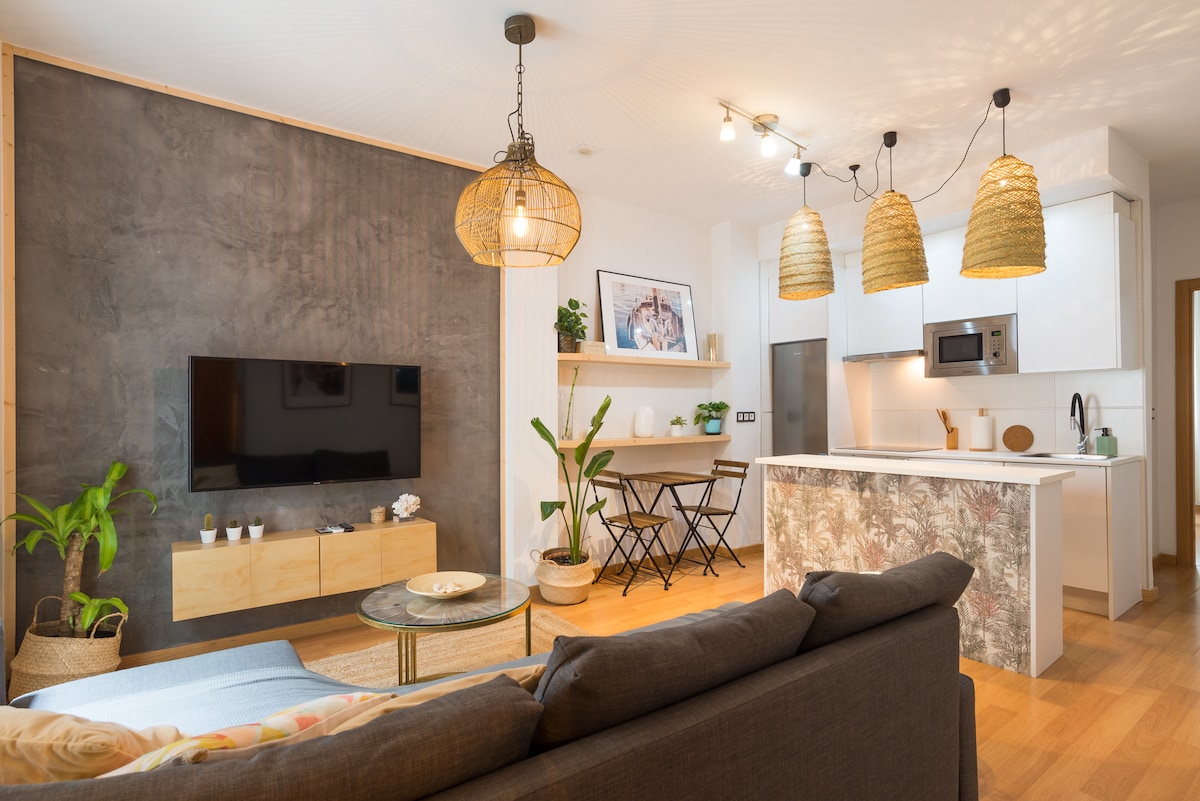 The living area features a comfortable sofa facing a wall-mounted TV. A dining area with two folding chairs is visible, accompanied by potted plants and a modern kitchen with sleek appliances. Soft lighting is provided by pendant lamps above the kitchen island.