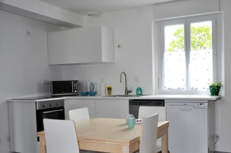 A modern kitchen features sleek white cabinetry and appliances, including a microwave, oven, and dishwasher. A small table with light-colored chairs is positioned in the center, while natural light filters through a window adorned with a sheer curtain, overlooking greenery outside.
