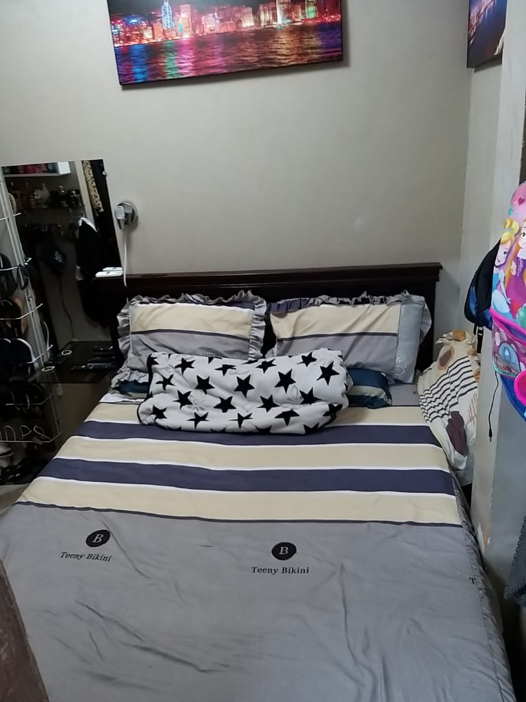 A small bedroom is presented, featuring a cozy bed adorned with striped linens in soft tones. Pillows with star patterns are arranged at the head of the bed, while a large, plush pillow adds comfort. A wall mirror reflects the space, contributing to the room's functionality.