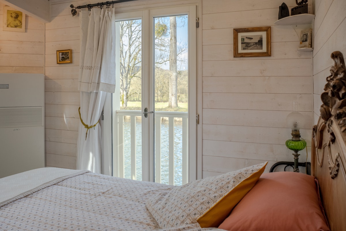 Natural light fills the room from the double doors, which open to a view of the water. Soft, neutral tones are present in the bedding, while decorative elements are placed on the walls and shelves, enhancing a serene atmosphere.