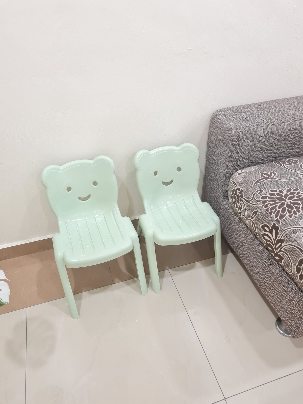 Two light green children's chairs with friendly face designs are positioned next to a textured brown sofa. The chairs are made of plastic and feature a simple, playful style. The soft, neutral tones of the surrounding area complement the cheerful seating.