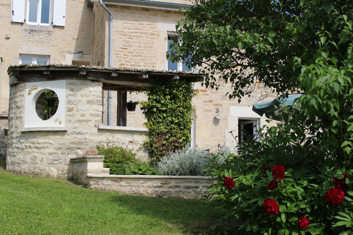 A charming stone exterior of a house is framed by greenery and seasonal flowers. A circular window is visible in a shaded alcove, complemented by a variety of plants. The well-maintained garden and lawn provide a peaceful outdoor space for relaxation.