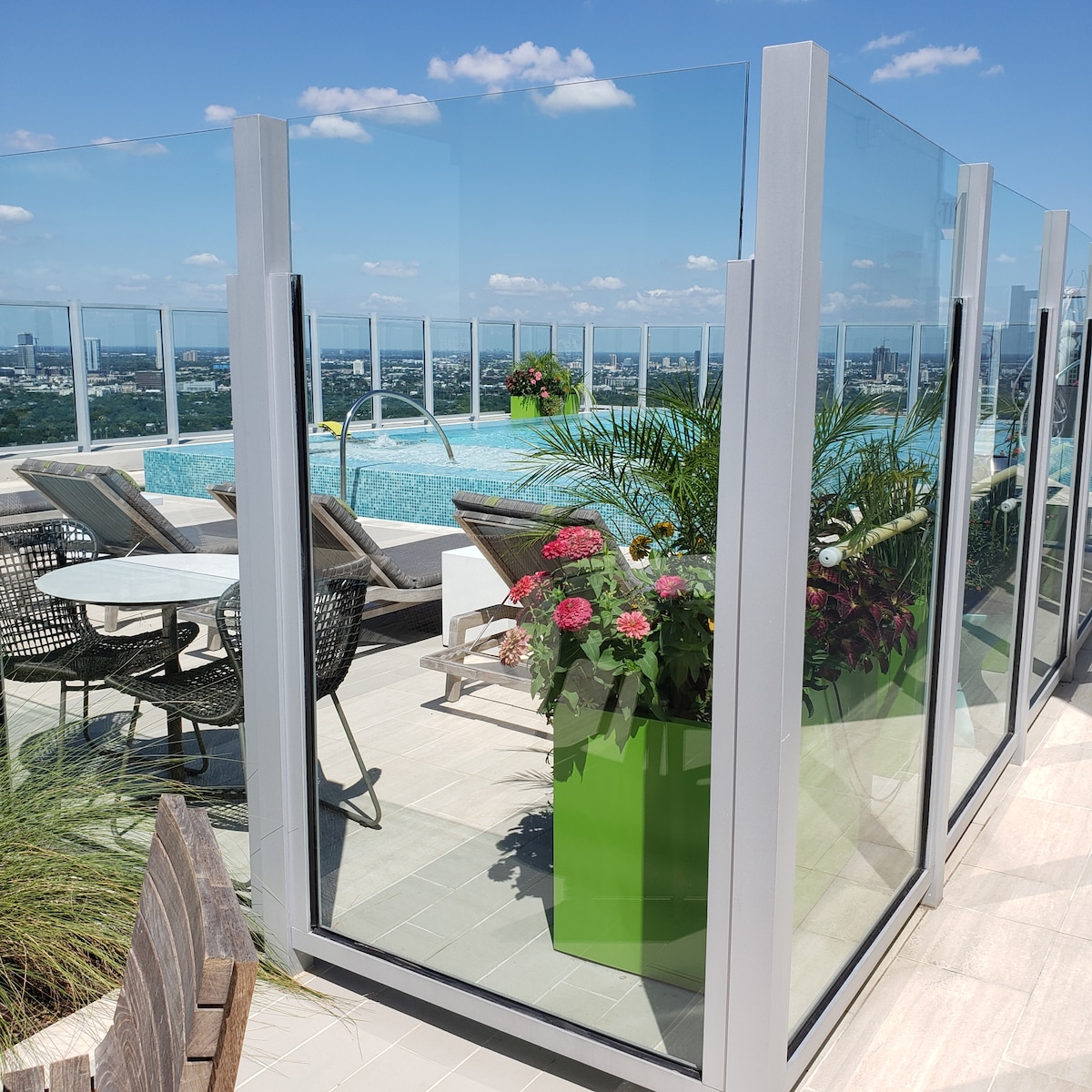 A rooftop pool area features glass paneling providing unobstructed views of the city. Lush greenery and vibrant potted flowers enhance the space, accompanied by comfortable seating options including lounge chairs and tables. A clear blue sky complements the tranquil atmosphere.