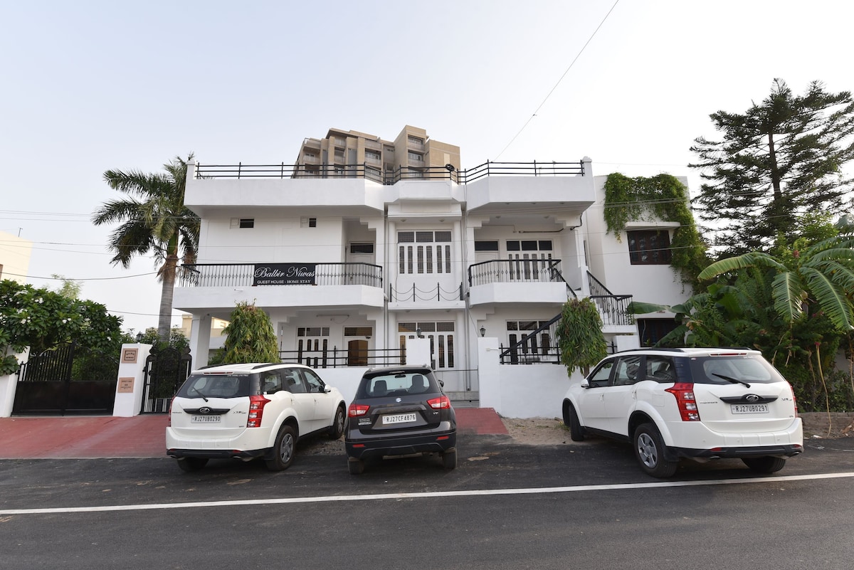 The exterior of a charming multi-story building is depicted, featuring a white facade with black accents and a second story balcony. Three parked vehicles line the driveway, with greenery enhancing the surroundings. Palm trees and shrubs are visible, contributing to a pleasant atmosphere.