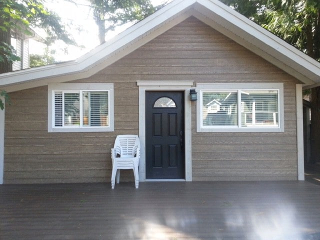 The exterior of the newly renovated cottage is shown, featuring a light brown wood-clad facade. Large windows allow natural light to enter, while a black front door provides a welcoming entrance. A white plastic chair is positioned beside the door on the wooden deck.