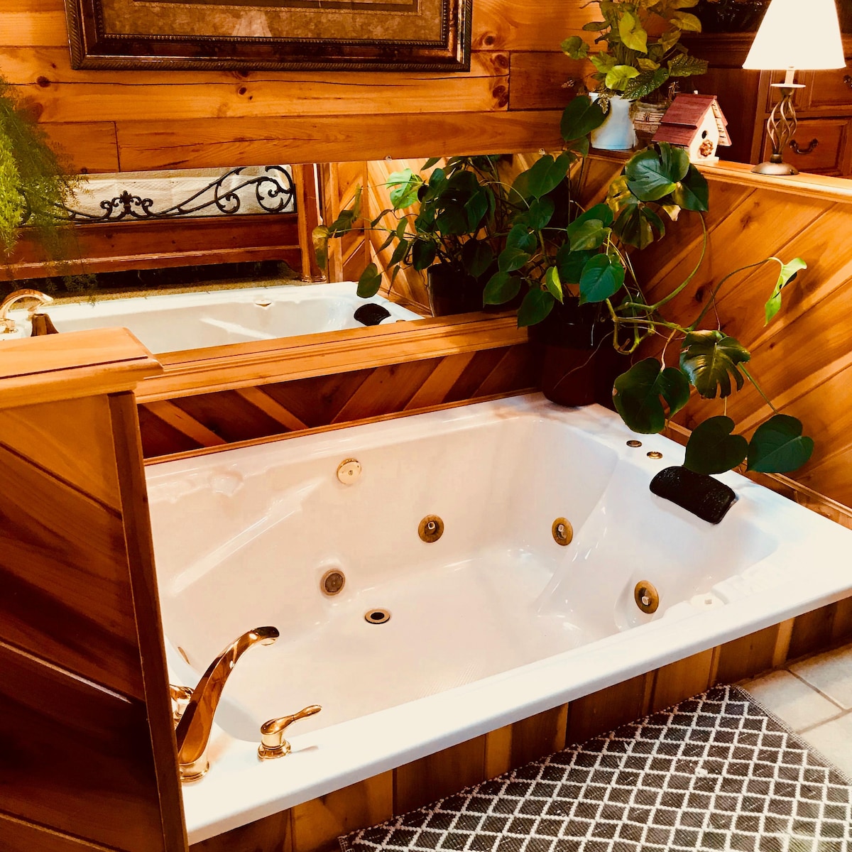 A spacious indoor Jacuzzi is set against a backdrop of warm wooden paneling, creating a cozy ambiance. Golden fixtures add a touch of elegance, while lush green plants provide a natural element. A soft rug lies beneath, enhancing the inviting environment.